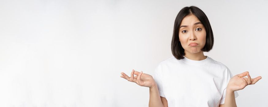 Portrait of confused asian woman shrugging shoulders, looking clueless, puzzled to say, standing over white background. Copy space