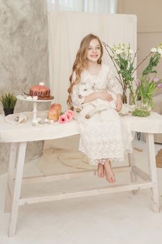 The girl is sitting at the Easter table with cakes, spring flowers and quail eggs and holding a white kid in her arms. Happy Easter celebration