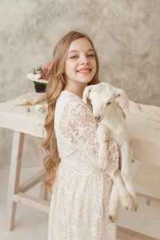 The girl is sitting at the Easter table with cakes, spring flowers and quail eggs and holding a white kid in her arms. Happy Easter celebration