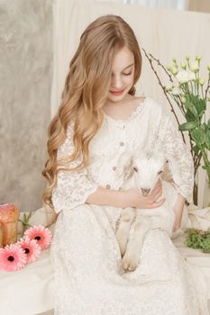 The girl is sitting at the Easter table with cakes, spring flowers and quail eggs and holding a white kid in her arms. Happy Easter celebration