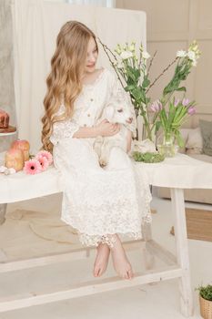 The girl is sitting at the Easter table with cakes, spring flowers and quail eggs and holding a white kid in her arms. Happy Easter celebration