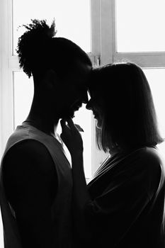 Black and white portrait, silhouette of a multiracial couple, boyfriend and girlfriend face to face, stand at the window. Vertical