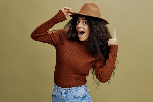 Great Idea concept. Overjoyed stylish curly Latin woman in brown hat, smiling at camera, holding hand on hat, pointing up, isolated green wall. People emotions lifestyle concept. Mock up copy space.