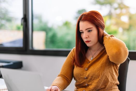 Young red-haired woman teleworking from home in a bright room with a large window behind. She is wearing a yellow sweater and is typing on the computer. The table is white with various desk items and lots of natural light. She is stroking her neck as she works.