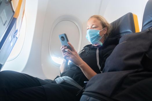 A young woman wearing face mask, using smart phone while traveling on airplane. New normal travel after covid-19 pandemic concept