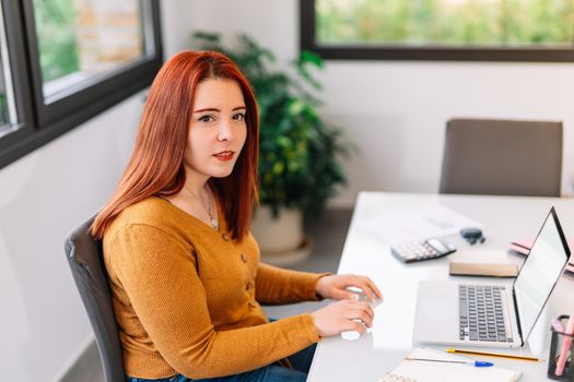 Young red-haired woman in yellow jumper looking at camera working from home with her laptop. Portrait of an enterprising and self-employed young girl teleworking at home with large window in the background, white table and plenty of natural light. Very clear image