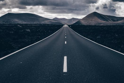 Endless road on a volcano in Timanfaya National Park in Lanzarote in the Canary Islands with a continuous line, black volcanic rocks on the side and volcanoes in mist in background