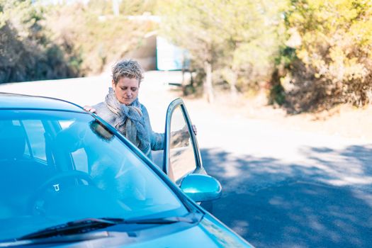 Modern adult mother entering her blue car through the gate on the road. She is on a small road next to a forest and mountain on a very sunny day. The girl is blonde and wearing long sleeves in grey and blue with a scarf around her neck.