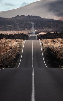 Endless road on a volcano in Timanfaya National Park in Lanzarote in the Canary Islands with a continuous line, black volcanic rocks on the side and volcanoes in mist in background.