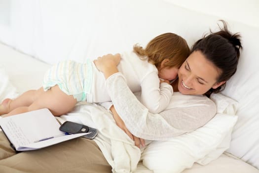 A young mother lying in bed and giving her toddler a hug with an open notebook lying on her back.