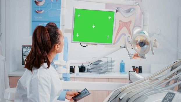 Dentist looking at monitor with horizontal green screen for oral care at dental office. Woman working as stomatologist with mockup template and isolated background for teethcare