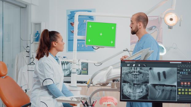 Dentist and assistant looking at green screen on monitor in dental cabinet. Teethcare specialists using chroma key and isolated mockup template for oral care, having dentistry tools