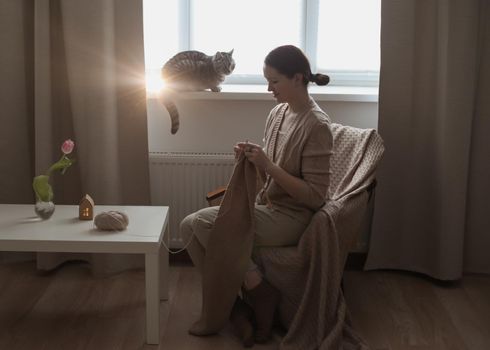 young woman knitting from woolen yarn and a cute funny tabby cat in cozy sunny room at home.