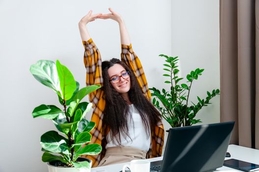 Smiling woman in glasses stretching on a workplace. Woman working at home using laptop computer sitting in living room.