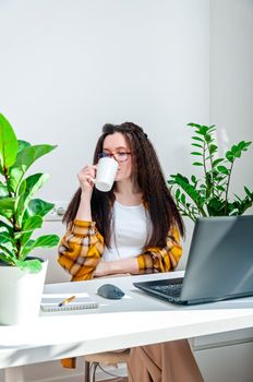 Beautiful woman in glasses drinks coffee on a break on workplace. Woman working at home, using laptop, undergoing training, watching webinar, online shopping.