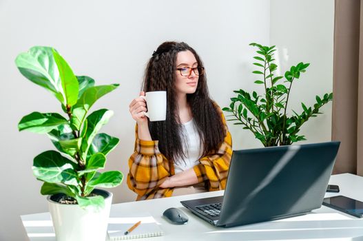 Beautiful woman in glasses drinks coffee on a break on workplace. Woman working at home, using laptop, undergoing training, watching webinar, online shopping.
