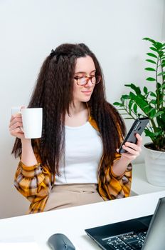 Beautiful woman in glasses drinks coffee on a break and using smartphone on workplace. Woman working at home, undergoing training, watching webinar, online shopping.