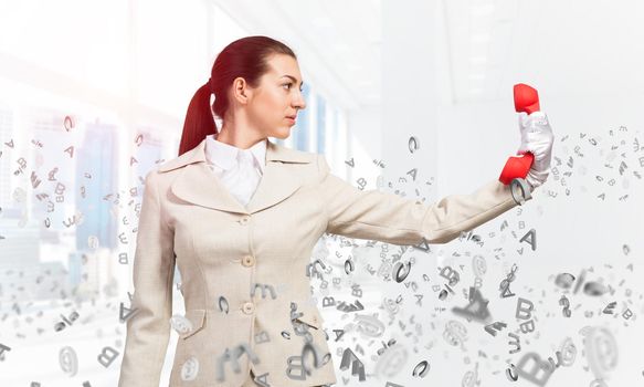 Woman keep at distance vintage red phone. Elegant operator in white business suit posing with landline phone in office with flying various letters. Hotline telemarketing and business communication.