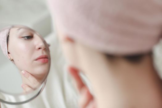 young woman with a towel on head looking in the mirror and apply cream on face skin.