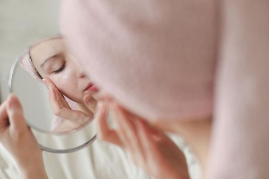 young woman with a towel on head looking in the mirror and apply cream on face skin.