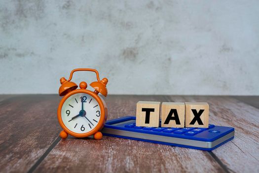 Tax text on Alphabet wooden block cube on a blue calculator with alarm clock set at 8 o'clock on wooden desk. Business and tax concept.