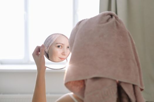 young woman with a towel on head looking in the mirror and apply cream on face skin.