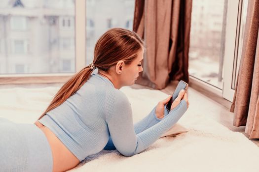 Side view portrait of relaxed woman with long hair lying on carpet at home. She is dressed in a blue tracksuit, holding a phone in her hands