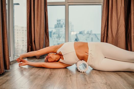 Adult athletic woman, in white bodysuit, performing fascia exercises on the floor - caucasian woman using a massage foam roller - a tool to relieve tension in the back and relieve muscle pain - the concept of physiotherapy and stretching training.