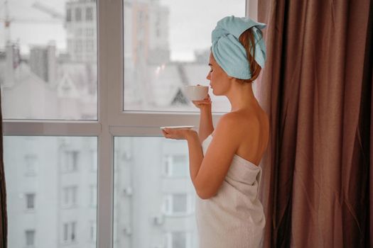 Young serene relaxed woman in spa bath towel drinking hot beverage tea coffee after taking shower bath at home. Beauty treatment, hydration concept