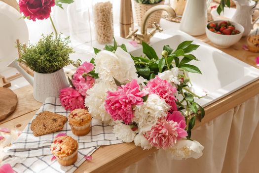 The kitchen countertop is decorated with peonies. The interior is decorated with spring flowers. Pink peonies and sweet cupcakes on a wooden countertop. Interior details