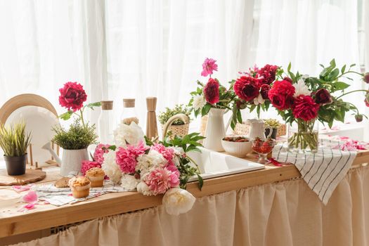 The kitchen countertop is decorated with peonies. The interior is decorated with spring flowers. Pink peonies and sweet cupcakes on a wooden countertop. Interior details