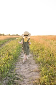 A little blonde girl in a straw hat walks in a field with a bouquet of daisies. The concept of walking in nature, freedom and an eco-friendly lifestyle