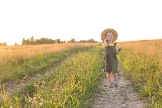 A little blonde girl in a straw hat walks in a field with a bouquet of daisies. The concept of walking in nature, freedom and an eco-friendly lifestyle