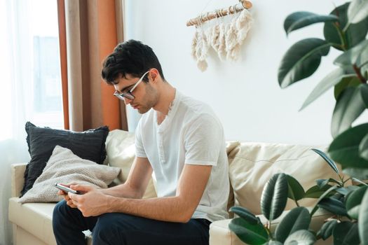 Attractive young man is using a tablet while sitting on the couch at home and browsing social networks and news