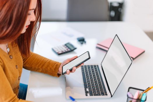 Pretty young red-haired girl in yellow sweater chatting on her smartphone while working on her laptop. Young businesswoman teleworking from home in a very bright environment with a large window in the background and natural light.