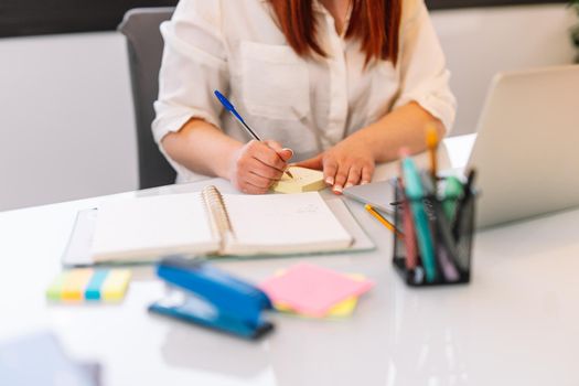 Hand shot of a young red-haired girl writing work matters in a notebook while consulting her laptop computer. Large white table with a white laptop, post-its of various colours yellow, pink and a jar with various pens and markers. Natural light illumination