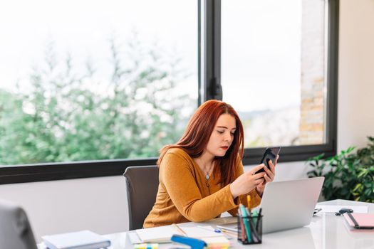 Pretty young red-haired girl in yellow sweater chatting on her smartphone while working on her laptop. Young businesswoman teleworking from home in a very bright environment with a large window in the background and natural light.