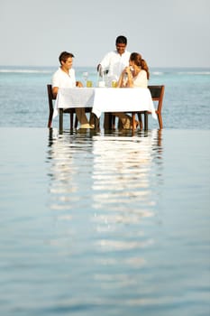 A young couple sitting at a table outside surrounded by water and being served wine.