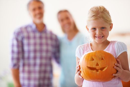 Portrait of a little girl holding her jack-o-lantern with her parents blurred behind her.