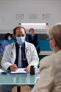 Doctor with face mask taking notes at checkup consultation, talking to senior woman in office. Specialist having conversation about medicine and health care with patient during pandemic.