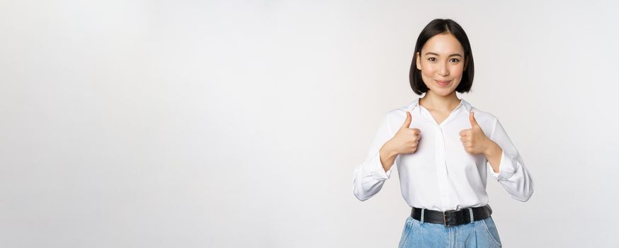 Image of beautiful adult asian woman showing thumbs up, wearing formal office, university clothing, recommending company, standing over white background.