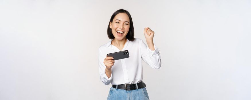 Happy asian woman do winner dance, triumphing, winning on mobile phone video game, holding smartphone horizontal position and celebrating, white background.