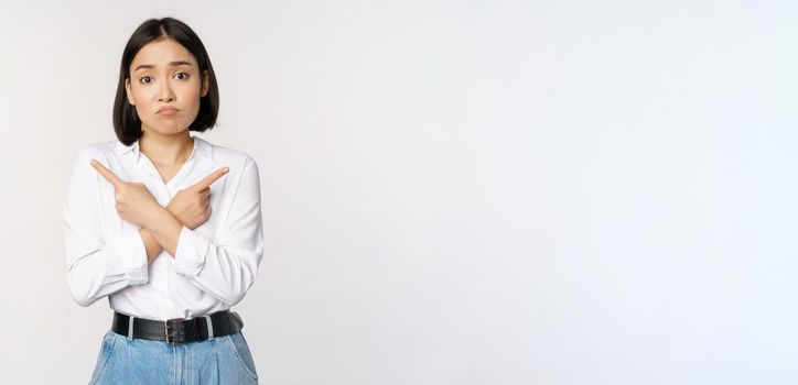 Confused sad asian girl cant decide, pointing fingers left and right, sulking and shrugging indecisive, cannot choose, standing over white background.