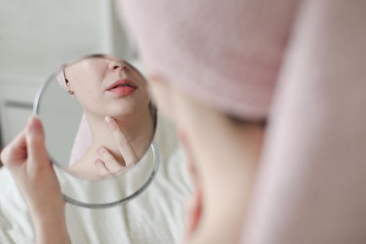 face of young beautiful healthy woman and reflection in the mirror. Young woman with a towel on head apply cream on face skin and doing make-up at home.