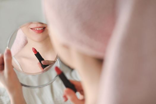 Beautiful attractive happy smiling woman paints lips with red lipstick using a small round mirror during home makeup in the morning.