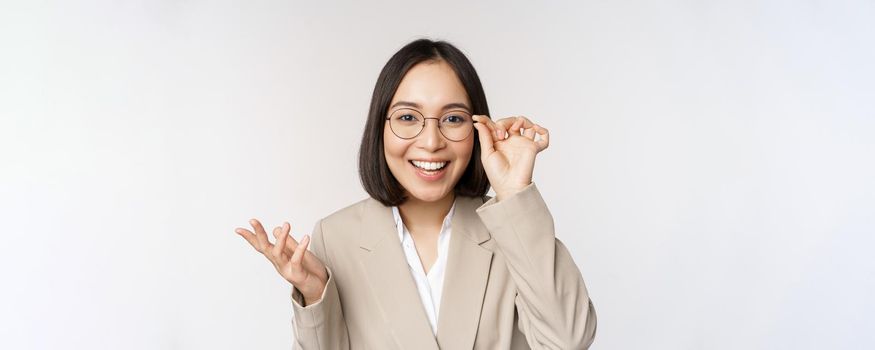 Enthusiastic asian saleswoman in glasses, smiling and laughing, looking amazed at camera, standing in beige suit over white background.