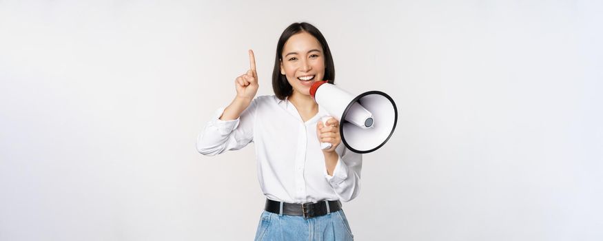 Smiling happy asian girl talking in megaphone and pointing up, announcing discount promo, showing advertisement on top, standing over white background.