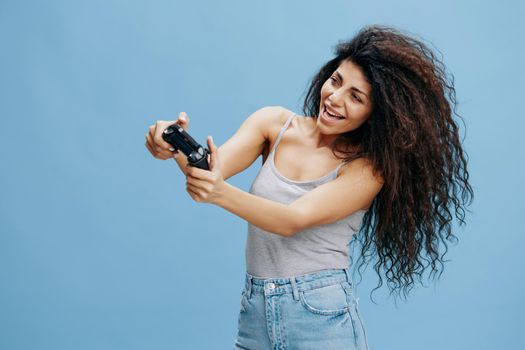 ONLINE GAME AD CONCEPT. Overjoyed beautiful curly Latin lady hold game pad near face, play cool fighting, look at side. Gaming accessories brands ad. Studio shoot isolated blue background