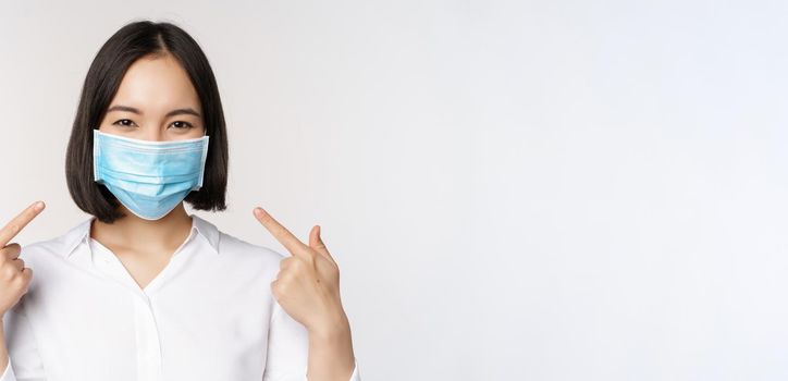 Image of young asian woman pointing at herself while wearing medical face mask, concept of covid-19 protection, standing over white background.