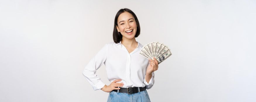 Credit and loan concept. Smiling young asian woman holding cash dollars and looking happy at camera, white background.